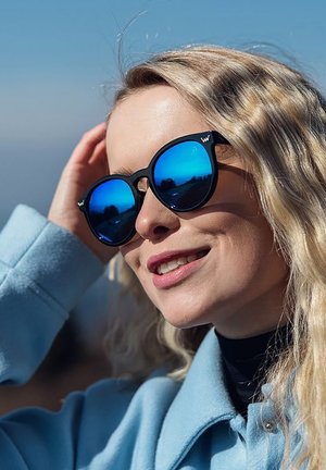 Černé sluneční brýle s modrými zrcadlovými čočkami, kulatý tvar a výrazné rámy. Vlnité blond vlasy, světle modrý kabát se stojáčkem.