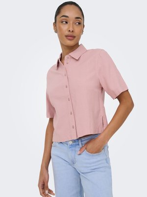 Kvinde iført en lyserød kortærmet skjorte med knapper og lyseblå jeans, med den ene hånd i lommen, der kigger sidelæns.