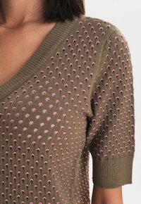 Nära bild av en olivgrön stickad topp med upphöjd rosa prickstruktur, ribbat V-hals och korta ärmar, bärs av en person.