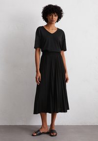 A young woman stands confidently in a black V-neck shirt and pleated black skirt, paired with simple black slide sandals against a neutral background.