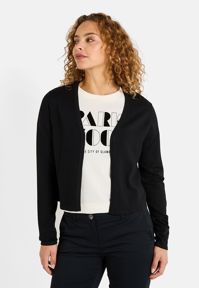 Cardigan noir en tricot à manches longues et à ouverture devant. Porté sur un t-shirt blanc avec un texte noir en gras. Texture lisse, coupe décontractée.
