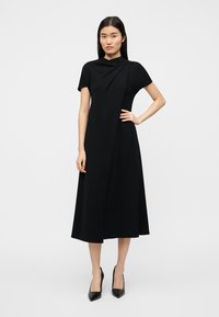 Μαύρο μίντι φόρεμα με κοντά μανίκια, απαλή υφή, ασύμμετρο σχέδιο τύλιγμα και διαγώνιο πλισέ λεπτομέρεια, συνδυασμένο με μαύρες μυτερές γόβες.