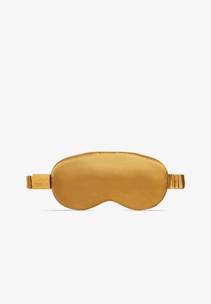 Donna sdraiata su un cuscino viola che indossa un pigiama beige, solleva una maschera per dormire in raso dorato dalla fronte.