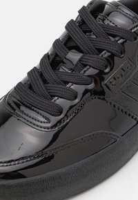 Gros plan d'une basket noire brillante avec des lacets noirs épais et une semelle texturée, présentant un logo Polo cousu sur le côté.