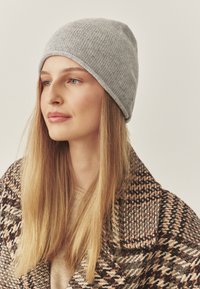 Gorro de punto gris con un diseño ajustado. El modelo lleva un abrigo con estampado, mostrando su cabello largo y lacio y una camiseta neutra debajo.