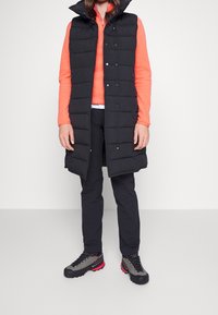 Svart pufferväst med hög krage, knäppt framtill, över en orange topp, i kombination med svarta byxor och grå vandringsskor med röda accenter.