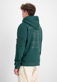 Zelena dukserica s kapuljačom i velikim grafičkim printom na leđima. Ima zippered džep na lijevoj rukavu i rebraste manšete.