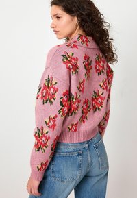 Vrouw in een roze gebreid sweater met rode en groene bloemmotieven en hoge taille blauwe jeans, die zijwaarts tegen een witte achtergrond staat.