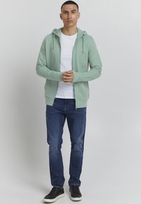 Mintgroene zip-up hoodie van zachte stof, over een wit T-shirt, in combinatie met donkerblauwe jeans en zwarte sneakers. Minimalistisch ontwerp.