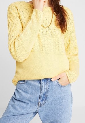 Personne portant un pull en maille torsadée jaune clair et un jean taille haute bleu clair, une main dans la poche, sur un fond uni.