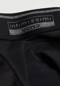 Svart tygmidja med en slät yta, med en grå etikett där "intimissimi" och skötselråd är tryckta.