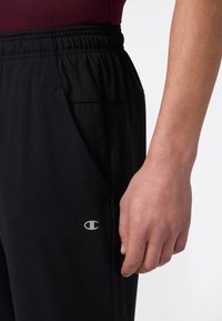 Close-up van een hand van een persoon naast zwarte elastische broeken met een klein Champion-logo nabij de zak, met een bordeauxrood topje.