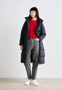 Abrigo acolchado largo negro con cuello alto, un suéter de punto rojo, jeans gris de cintura alta y zapatillas blancas, de pie sobre un fondo neutro.