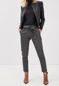 Kvinna som bär grå högmidjade byxor med frontknut, svart ribbad polotröja, svart läderjacka och bruna mocka ankelstövlar.