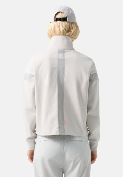 Person with short blonde hair wearing a light gray high-collar jacket and matching pants, with a white cap worn backwards.