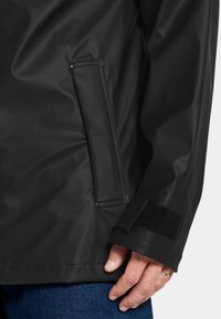 Svart vattentät jacka med en slätt yta, sidoficka med klaffstängning och justerbar kardborrekant på ärmarna. Tyget ser hållbart ut.