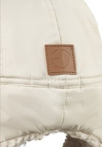 Beige isoleret hat med tekstureret ydre materiale, syede detaljer og et brunt lædermærke med et rundt logo. Fleeceforing er synlig.