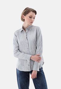 Light gray button-up shirt made of linen, featuring textured floral patterns. Long sleeves with button cuffs, relaxed fit. Dark jeans visible.