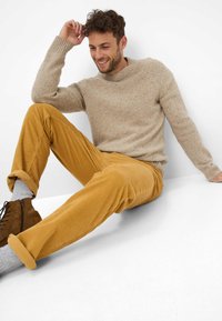 Beige gebreide trui met geribbelde manchetten, gecombineerd met mosterdkleurige corduroy broek. Accessoires zijn grijze sokken en bruine schoenen met veters.