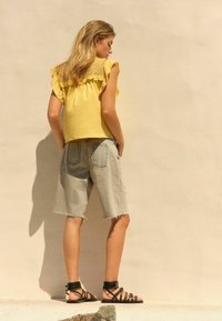 Gul volangtop med spetsbak, ljusblå denimshorts och svarta sandaler med remmar. Outfiten har en avslappnad och ledig stil.