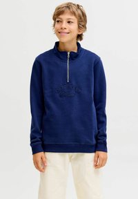 Námořnická modrá mikina na zip s vysokým límcem a žebrovanými manžetami, s vyraženým nápisem "ORIGINALS" na přední straně. Nošena s béžovými kalhotami.