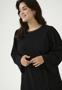 Vestido negro con una tela texturizada, que presenta un escote festoneado y mangas acampanadas. Los detalles laterales añaden interés visual.
