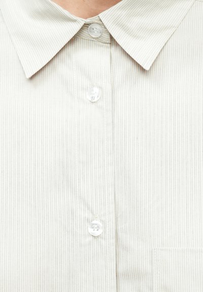 Világos bézs gombos ing vékony függőleges csíkokkal, klasszikus gallérral és egyetlen mellzsebbel. Sima textúra és műanyag gombok.