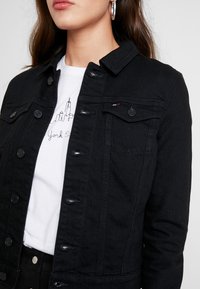 Woman wearing silver hoop earrings, black denim jacket, and white T-shirt with minimalist New York City skyline and handwritten text.