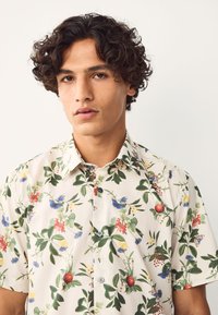 Camicia a maniche corte con motivo floreale su sfondo crema, con vari motivi colorati di fiori e frutti, e colletto con bottoni.