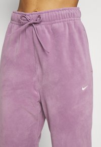 Lila Fleecejogginghosen mit elastischem Bund, Tunnelzug und kleinem weißen Nike-Logo am linken Oberschenkel. Weiche Textur.