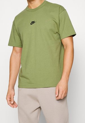 Homme portant un t-shirt Nike vert olive avec un logo noir et un pantalon de jogging beige clair, debout devant un fond blanc.