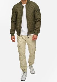 Olivgrüne gesteppte Bomberjacke mit gerippten Bündchen, seitlicher Reißverschlusstasche, getragen über einem weißen T-Shirt und beigen Cargo-Hosen mit Taschen.