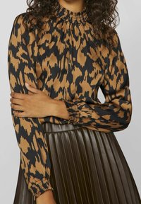 Blusa con stampa animale marrone e nera, dal collo alto e gathered e maniche lunghe a forma di palloncino; il tessuto ha una consistenza liscia e lucida.