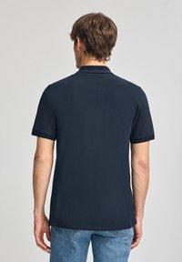 Camisa polo de color azul marino con mangas cortas, cuello acanalado y textura de algodón suave. Llevada con jeans de color azul claro, mostrando un ajuste casual.