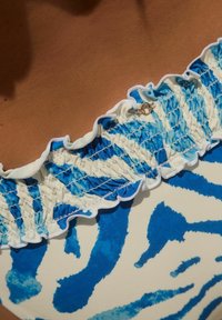 Blauw en witte eendelig badpak met een geplooide halslijn met gerimpelde randen en een kleine gouden accentdetail. De stof heeft een aquarelpatroon.