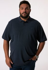 Tmavě modré polo tričko z prodyšného materiálu, s límečkem, plaketou na tři knoflíky a nenápadným logem. Spárováno s tmavými džínami.