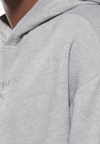 Gros plan sur une personne portant un sweat à capuche gris clair texturé avec un logo brodé subtil sur la poitrine.