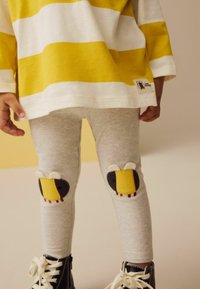 Graue Leggings mit Bienenapplikationen an den Knien, kombiniert mit einem gelb-weiß gestreiften Langarmshirt. Schwarze Schuhe runden das Outfit ab.