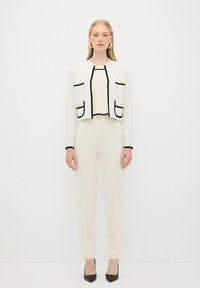 Veste blanche courte avec des bordures noires et des accents dorés, associée à un haut crème assorti et un pantalon ajusté. Des escarpins noirs à talons pointus complètent le look.