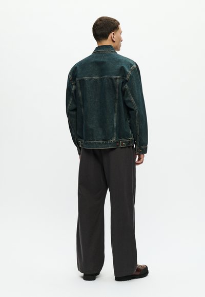 Uomo di spalle che indossa una giacca di jeans blu scuro, pantaloni larghi scuri e scarpe marroni su uno sfondo bianco uniforme.