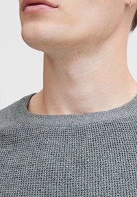 Grå tekstureret striktrøje med rund halsudskæring. Bemærkelsesværdig ribbet detalje ved ærmerne og halsen. Nærbillede som fokuserer på den øverste del af overkroppen.