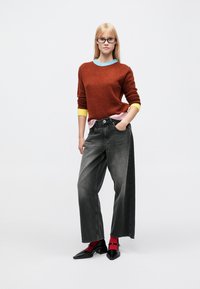 Pullover in Rostfarbe mit gelben und blauen Akzenten, kombiniert mit weiten, ausgeblichenen schwarzen Jeans. Schwarze Schuhe und rote Socken vervollständigen das Outfit.