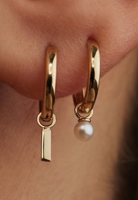 Boucles d'oreilles en or avec un pendentif en barre verticale et une seule perle blanche, mettant en avant une finition polie et une texture lisse.