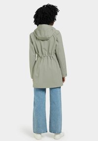 Veste de pluie à capuche vert clair, avec une taille élastique à cordon de serrage, portée sur un jean large bleu et des baskets blanches. Texture lisse.