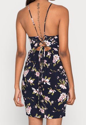 Nainen pukeutunut tummansiniseen kukkakuosiseen mekkoon, jossa on avoin selkä ja ristikkäin solmitut hihnat alaselässä, paljastaen pystysuoran tatuoinnin selkärangallaan.