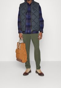 Wytłaczana ciemnozielona kamizelka na niebieskiej koszuli w kratę, oliwkowe spodnie, brązowe i pomarańczowe buty oraz beżowa torba na ramię z klapą.