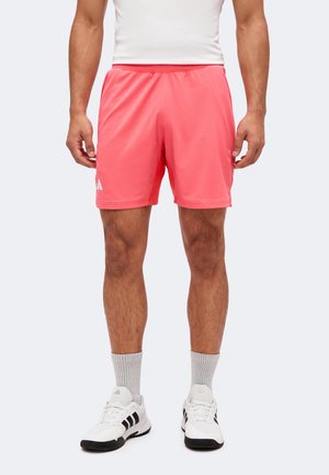 Homme portant un short de sport corail, des chaussettes blanches montantes et des chaussures de sport blanches avec des rayures noires, debout devant un fond clair uni.