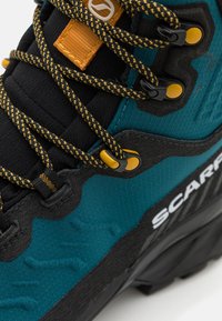 Botte de randonnée teal et noire avec un matériau texturé, des lacets jaunes et noirs, et une étiquette d'accent orange ; possède une semelle robuste pour une meilleure adhérence.