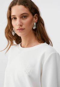 Sweatshirt blanc à encolure ronde, avec un logo brodé discret sur la poitrine. Le modèle porte de grandes boucles d'oreilles métalliques.