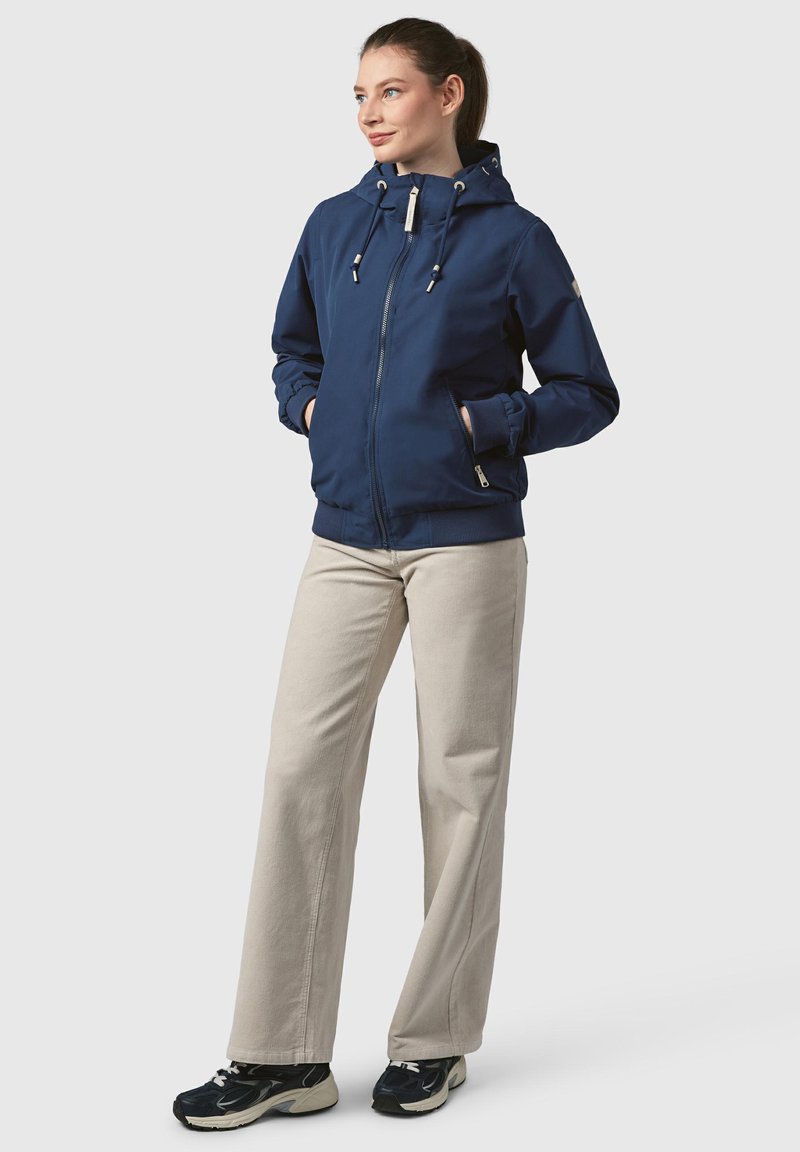 Marinblå zip-up jacka med huva, dragskor och sidofickor. Parades med ljusbeiga byxor och svarta sneakers. Minimalistisk design.